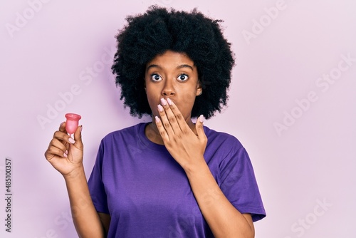 Young african american woman holding menstrual cup covering mouth with hand  shocked and afraid for mistake. surprised expression