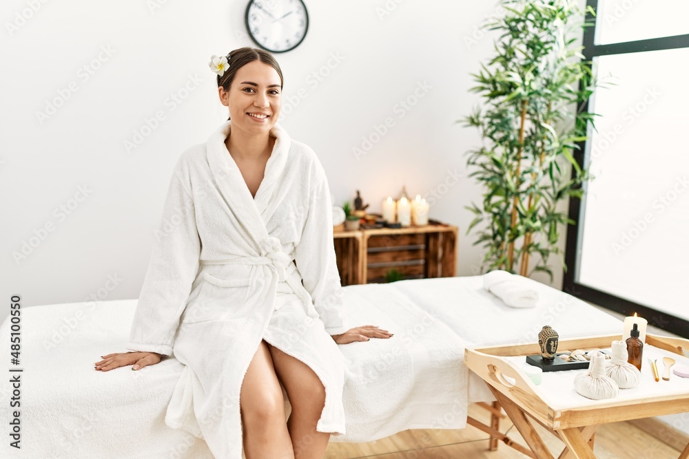 Young hispanic woman smiling confident sitting on massage table at beauty center.