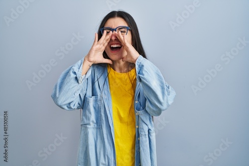Young hispanic woman standing over blue background shouting angry out loud with hands over mouth