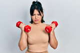 Young hispanic woman wearing sportswear using dumbbells skeptic and nervous, frowning upset because of problem. negative person.