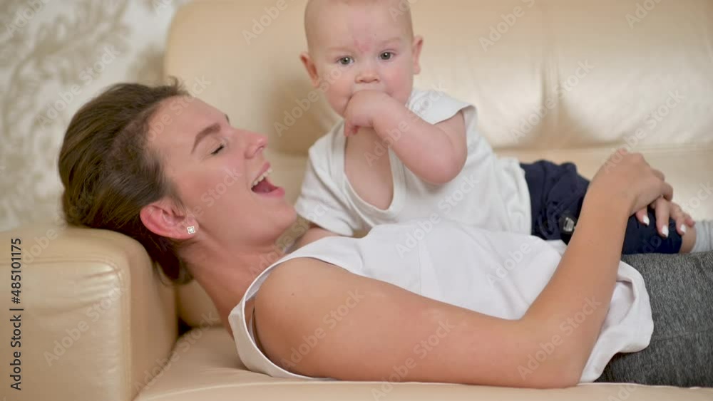 Young happy mother is playing with her newborn baby on the sofa in the morning. smiling mum enjoying bonding cuddling hugging spending time with baby. Parenthood, Childhood. Young happy mother is playing with her newborn baby on the sofa in the morning. smiling mum enjoying bonding cuddling hugging spending time with baby. Parenthood, Childhood.