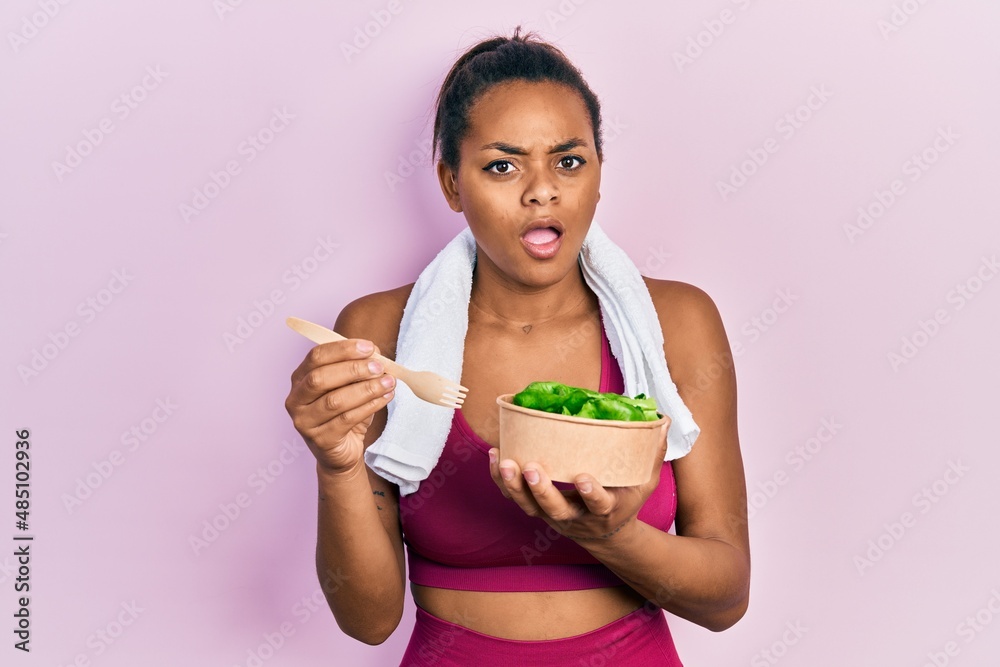 Young african american girl wearing sportswear eating salad in shock face, looking skeptical and sarcastic, surprised with open mouth