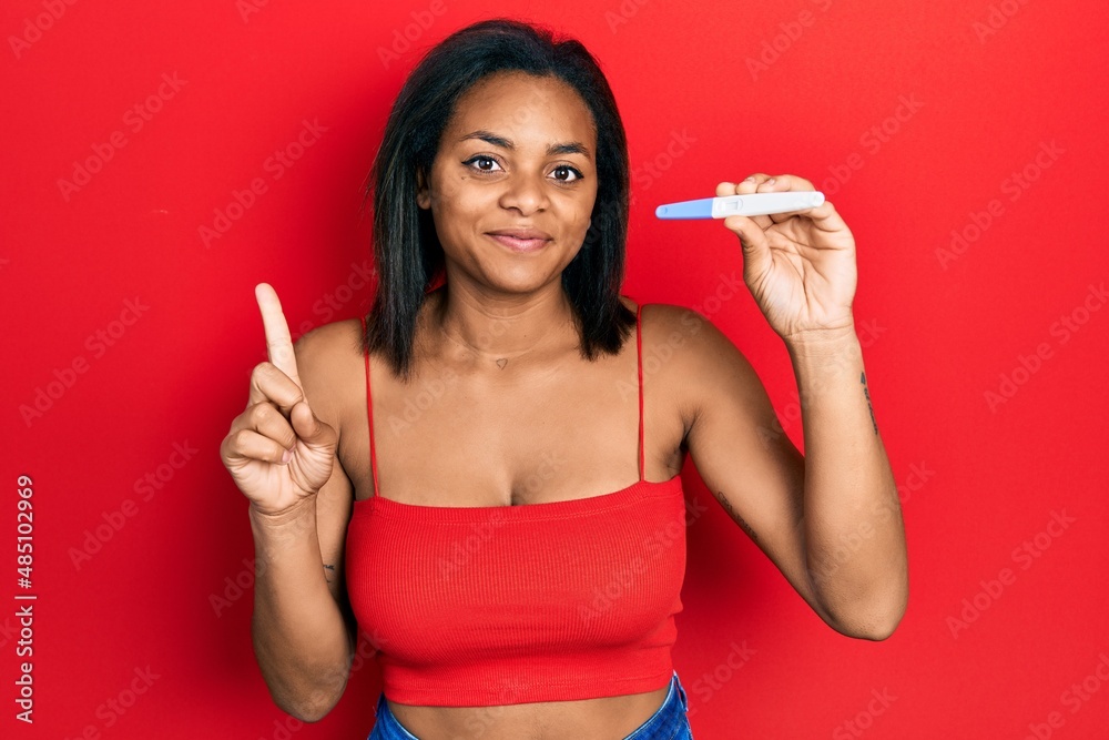 Young african american girl holding pregnancy test result smiling with an idea or question pointing finger with happy face, number one Young african american girl holding pregnancy test result smiling with an idea or question pointing finger with happy face, number one