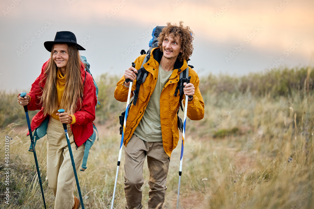 excited couple with bag pack on back holding hiking pole during hiking jurney, happy athletic man and woman in colorful sportive outfit, vacation in nature outdoors. travel, sport, adventure concept