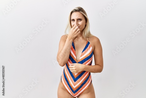 Young blonde woman wearing swimsuit over isolated background smelling something stinky and disgusting, intolerable smell, holding breath with fingers on nose. bad smell