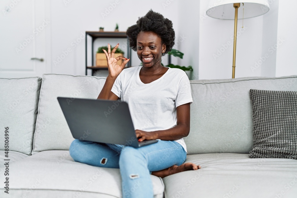 African young woman using laptop at home doing ok sign with fingers, smiling friendly gesturing excellent symbol