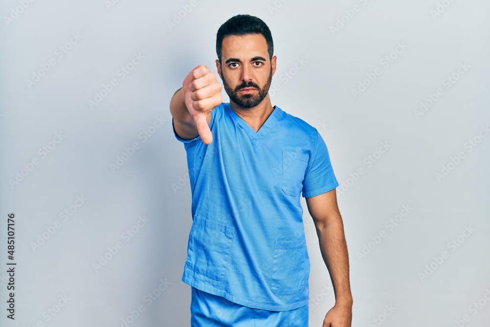 Handsome hispanic man with beard wearing blue male nurse uniform looking unhappy and angry showing rejection and negative with thumbs down gesture. bad expression. Handsome hispanic man with beard wearing blue male nurse uniform looking unhappy and angry showing rejection and negative with thumbs down gesture. bad expression.