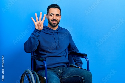 Handsome hispanic man with beard sitting on wheelchair showing and pointing up with fingers number four while smiling confident and happy.