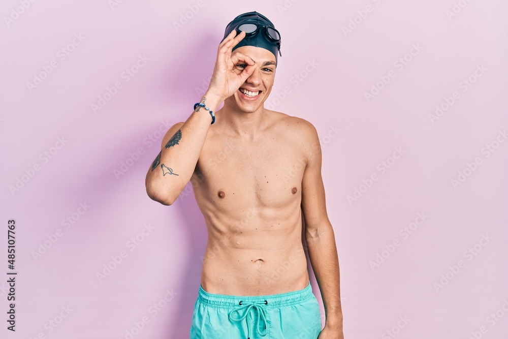 Young hispanic man wearing swimwear and swimmer glasses doing ok gesture with hand smiling, eye looking through fingers with happy face.