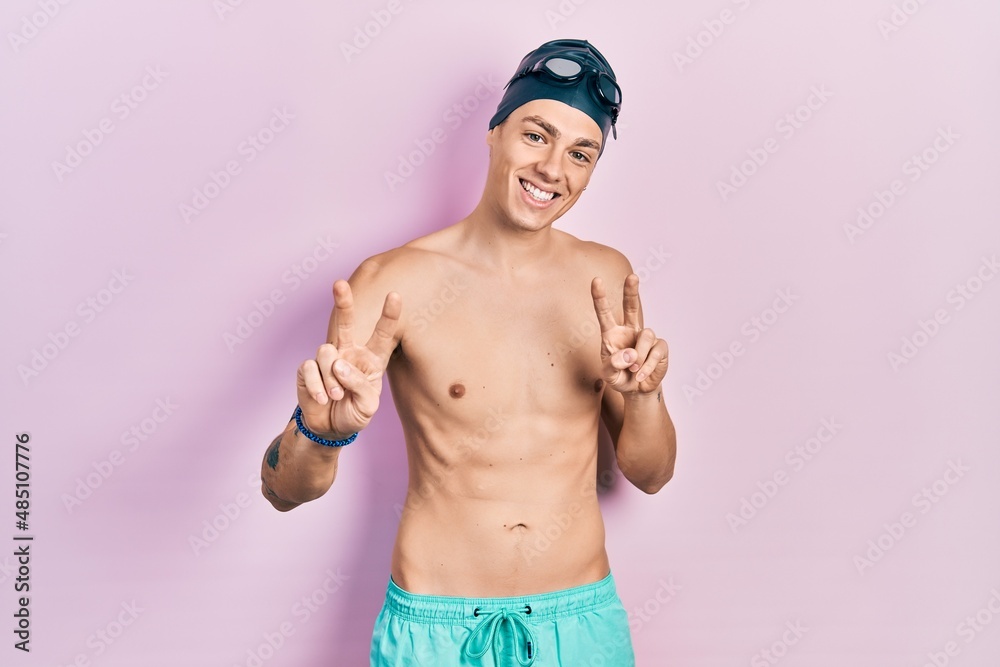 Young hispanic man wearing swimwear and swimmer glasses smiling looking to the camera showing fingers doing victory sign. number two.