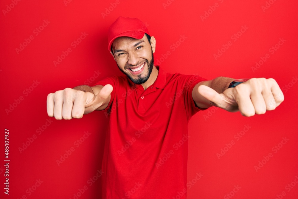 Hispanic man with beard wearing delivery uniform and cap approving doing positive gesture with hand, thumbs up smiling and happy for success. winner gesture. Hispanic man with beard wearing delivery uniform and cap approving doing positive gesture with hand, thumbs up smiling and happy for success. winner gesture.