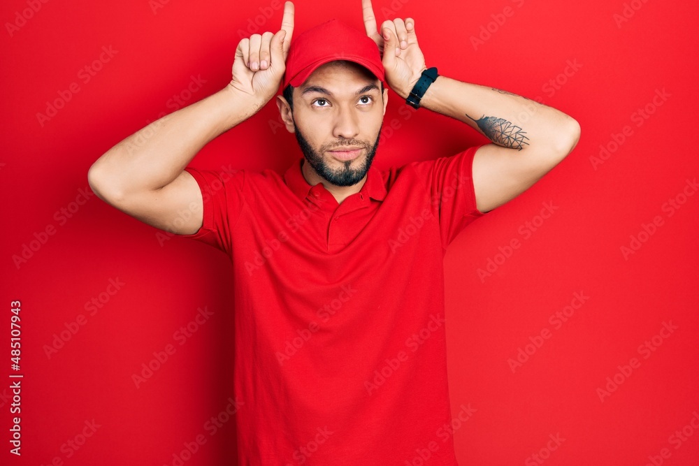 Hispanic man with beard wearing delivery uniform and cap doing funny gesture with finger over head as bull horns Hispanic man with beard wearing delivery uniform and cap doing funny gesture with finger over head as bull horns