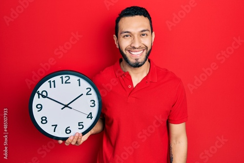 Hispanic man with beard holding big clock looking positive and happy standing and smiling with a confident smile showing teeth