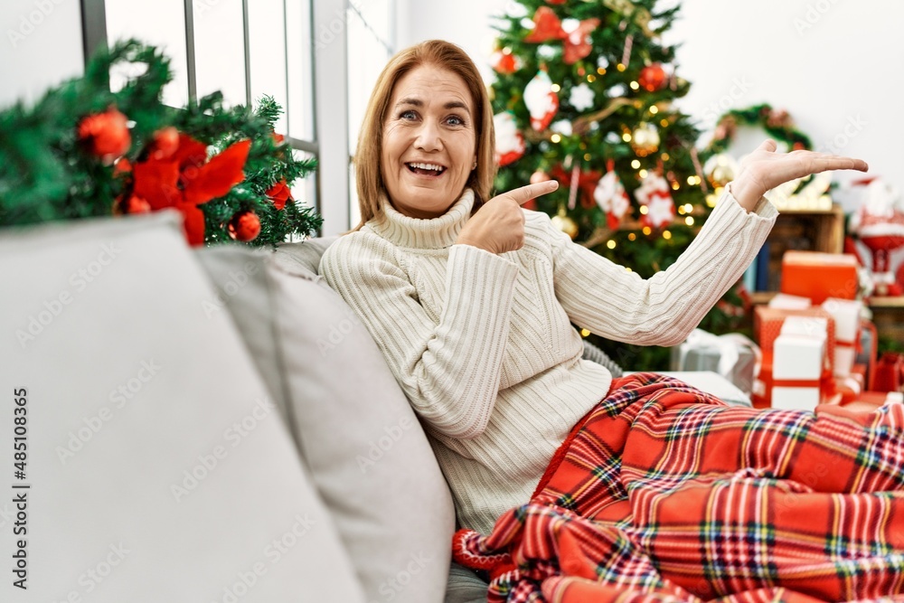 Middle age woman sitting on the sofa by christmas tree amazed and smiling to the camera while presenting with hand and pointing with finger.