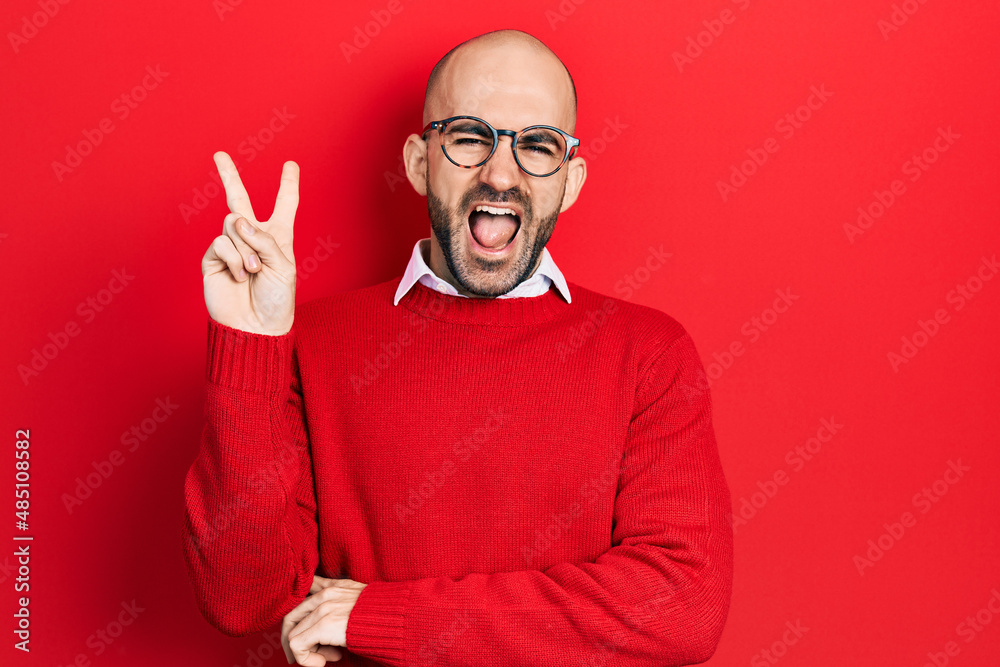 Naklejka premium Young bald man wearing casual clothes and glasses smiling with happy face winking at the camera doing victory sign. number two.