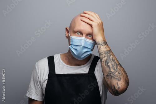 Surprised and frightened bald chef or butcher holding his head. On gray background.
