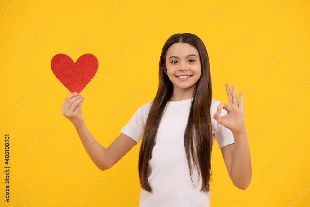 happy teen girl holding valentines heart showing ok gesture on yellow background, love happy teen girl holding valentines heart showing ok gesture on yellow background, love