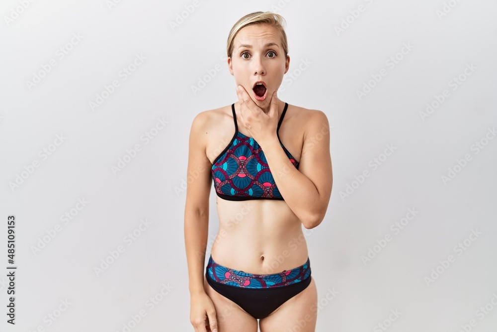 Naklejka premium Young blonde woman wearing sporty bikini over isolated background looking fascinated with disbelief, surprise and amazed expression with hands on chin