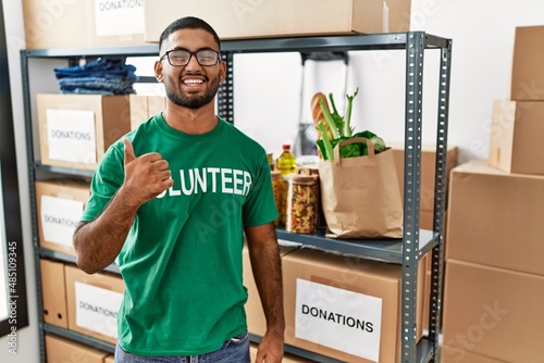 Young indian man volunteer holding donations box doing happy thumbs up gesture with hand. approving expression looking at the camera showing success.