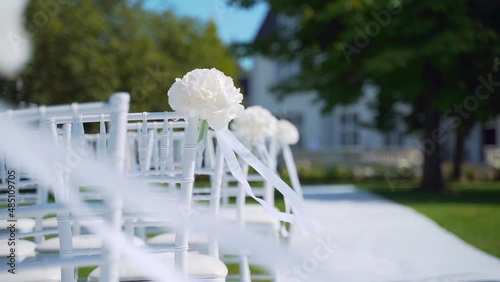 Close up. White chairs for guests at a luxury outdoor wedding ceremony. Event decoration with fresh flowers. Beautiful, convenient. armchair wind blows a ribbon on a lawn. Before celebration. Details