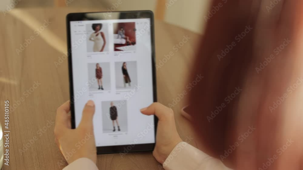 Rear view of female hand touching tablet screen while selecting dress on ecommerce portal. Lady use e-commerce webshop to buy dress. Modern devices, online, shopping concept