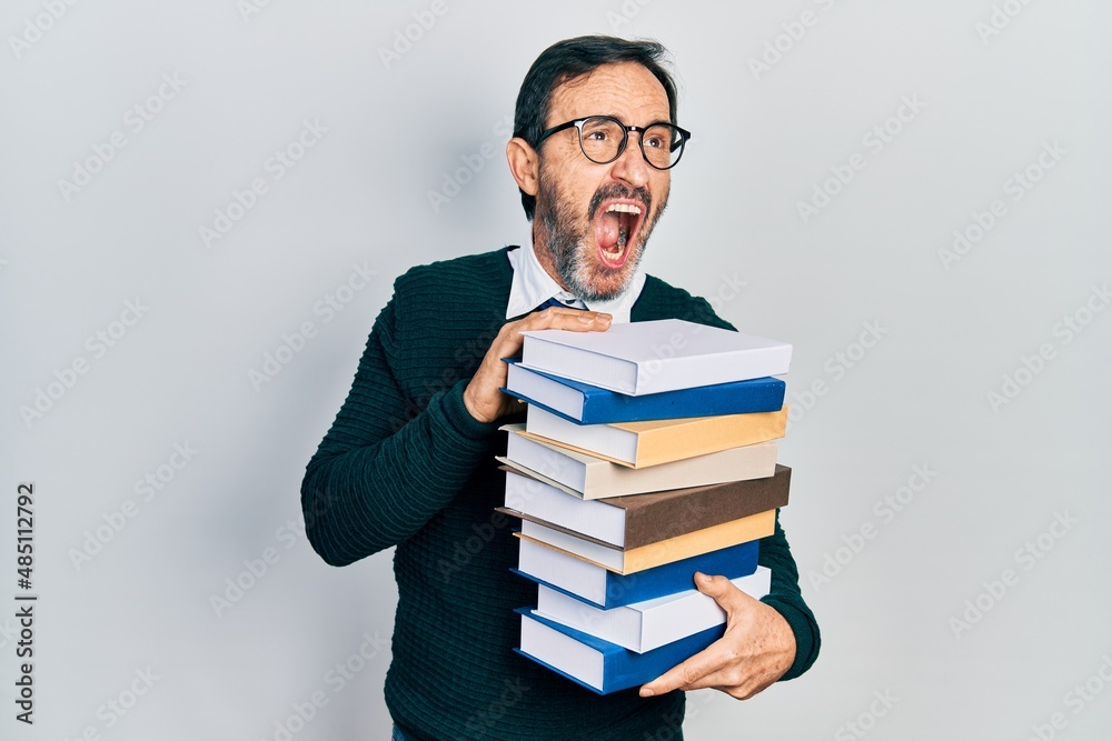 Middle age hispanic man holding a pile of books angry and mad screaming frustrated and furious, shouting with anger. rage and aggressive concept. Middle age hispanic man holding a pile of books angry and mad screaming frustrated and furious, shouting with anger. rage and aggressive concept.