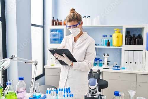 Young blonde woman wearing scientist uniform and medical mask writing on clipboard at laboratory