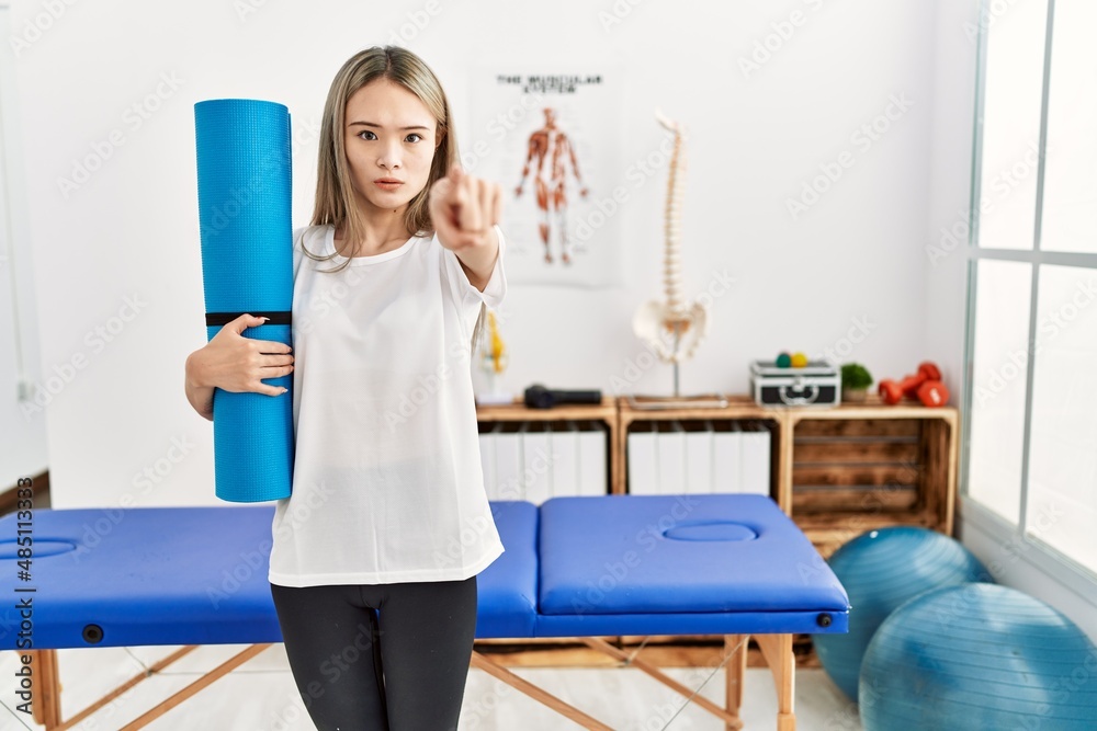 Asian young woman holding yoga mat at pain recovery clinic pointing with finger to the camera and to you, confident gesture looking serious