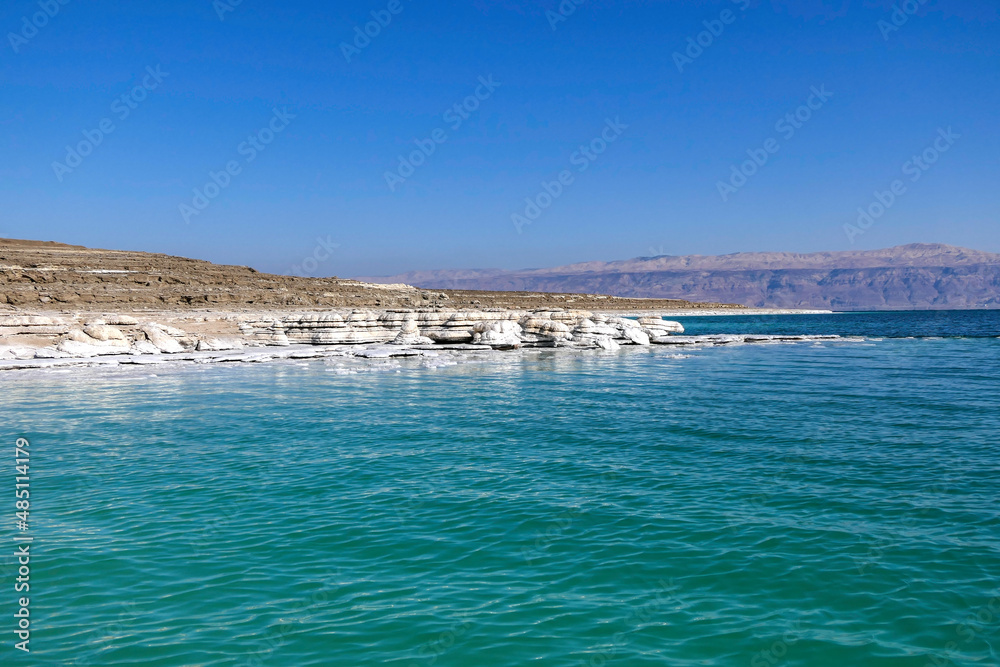 View of the mountains across the waters of the Dead Sea from the shore covered with salt formations.
