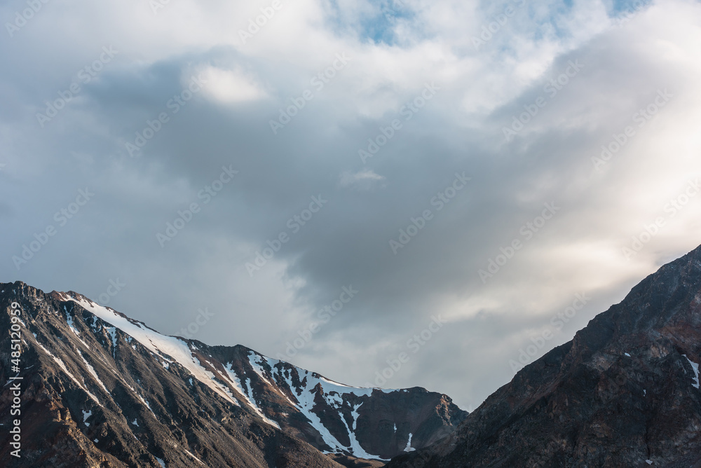 Dramatic overcast landscape with sunlit high mountain range with sharp rocks under gloomy sky. Dark atmospheric mountain scenery with large mountains in sunlight under cloudy sky at changeable weather