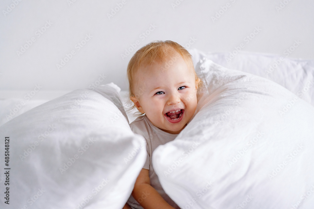 Laughing toddler having fun on bed with lots of pillows. A baby playing hide and seek.