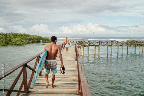 Hobby and vacation. Holiday on the beach. Back view of young man carrying surf board on wooden bridge. © luengo_ua