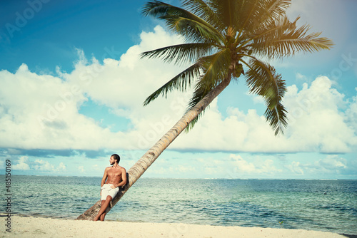 Enjoying suntan and vacation. Portrait of young bearded man leaning on coconut palm tree on the tropical beach.