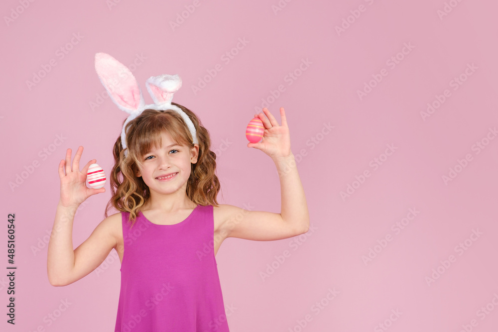 Closeup portrait of smiling little girl in Easter bunny ears smiling while holds in hands colorful eggs against pink background
