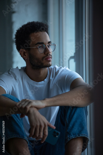 Attractive african american man in glasses and a white t-shirt sits on a windowsill.