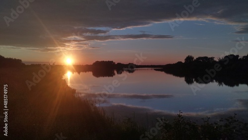 The sun is setting over the river among the clouds. The sky and clouds are reflected in the calm water. Tall grass and trees grow on the banks of the river