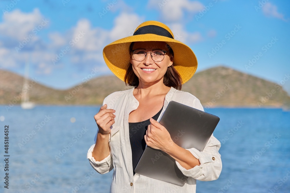 Portrait of happy mature woman in straw hat with laptop on the beach