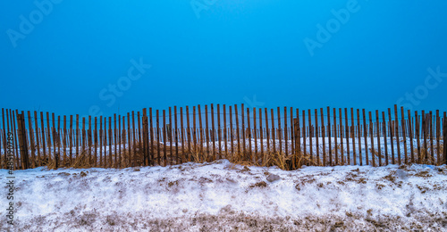 Clear blue sky, wooden snow fences, and snow piles on the coastal highway. Abstract geometry of the vertical fence pieces, horizontal snow pile, and plain turquoise-colored ocean background.