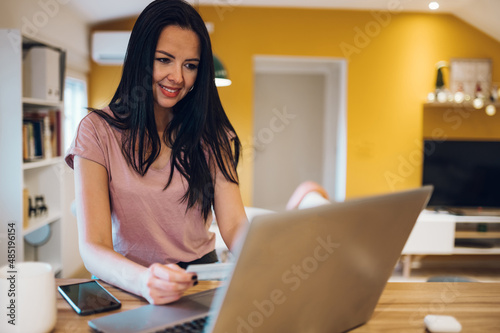 Middle aged woman using laptop and credit card while sitting at home kitchen