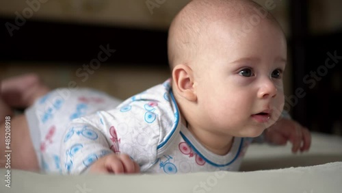 Newborn Active Baby Cute Smiling Teethless Face Portrait Early Days On Stomach Developing Neck Control. 5 Months Child On White Bed Looking on Camera. Infant, Childbirth, Parenthood, Beginning Concept photo