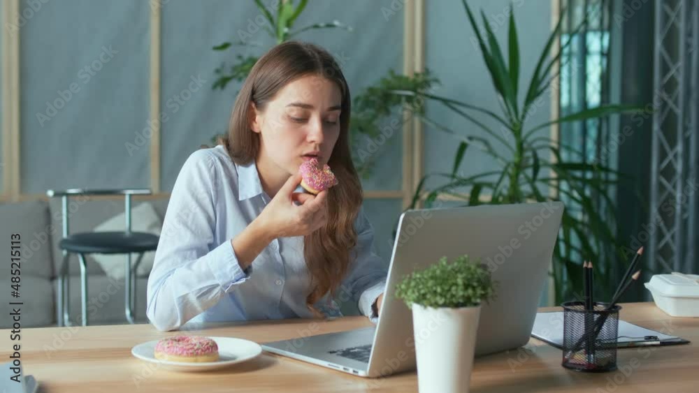 Young Businesswoman Eating a Glazed Donut While Working on a Laptop at the Office Table. A Quick Snack, High-calorie Food, Weight Problems, Diabetes Mellitus. Eating Disorder and Business Concept. Young Businesswoman Eating a Glazed Donut While Working on a Laptop at the Office Table. A Quick Snack, High-calorie Food, Weight Problems, Diabetes Mellitus. Eating Disorder and Business Concept.