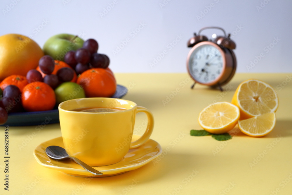 Healthy breakfast of fruits and tea with lemon. There is a yellow tablecloth on the table, a cup of tea is yellow. There is a blurry alarm clock in the photo that shows 7 o'clock. Healthy breakfast of fruits and tea with lemon. There is a yellow tablecloth on the table, a cup of tea is yellow. There is a blurry alarm clock in the photo that shows 7 o'clock.