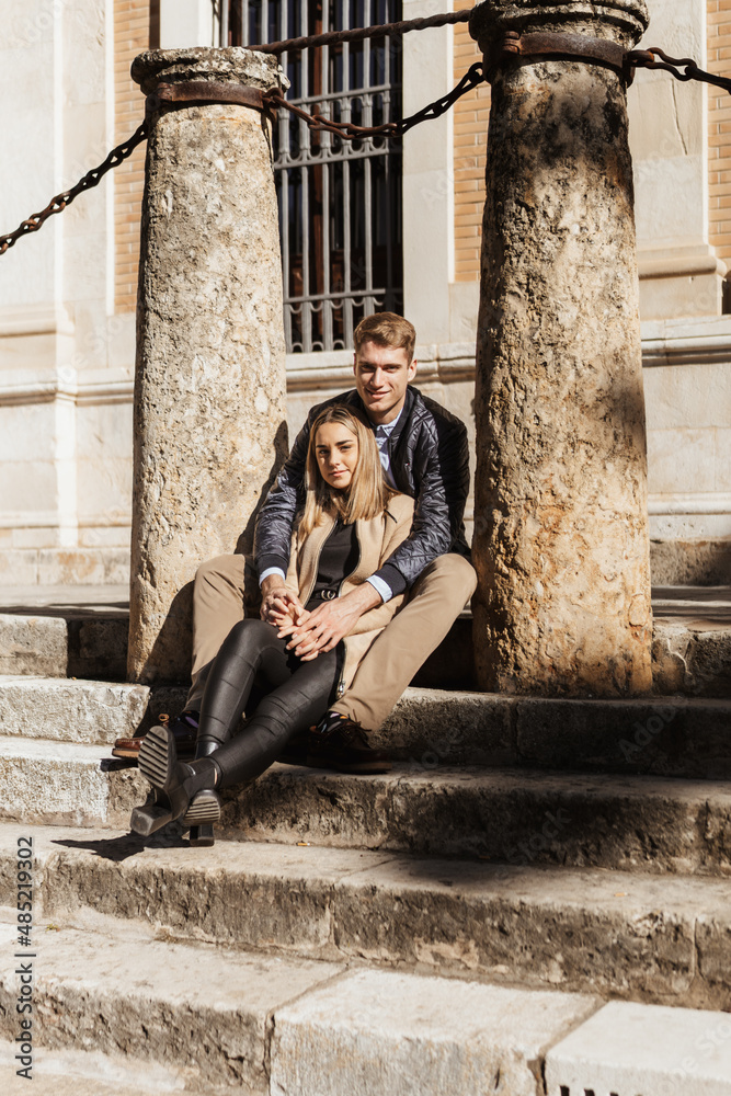 A couple sitting on a staircase and between two columns look at the camera.