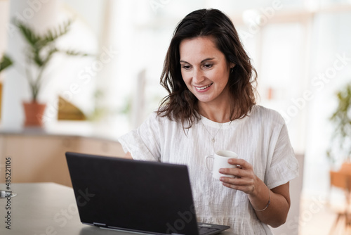 happy smiling remote working woman drinking coffee or espresso infront of a laptop or notebook on her work desk in her modern airy bright living room home office 