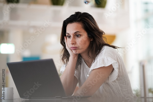 sad depressed thoughtful remote working brown hair woman sitting disappointed  infront of a laptop or notebook on her work desk in modern airy bright living room home office  