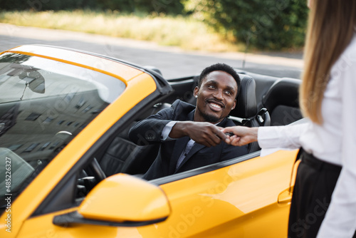 Young handsome African businessman in a Car Rental Service. Test Drive Concept. Young African American man sitting inside new yellow cabriolet car while taking keys from female seller.