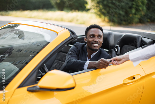 Young handsome African businessman in a Car Rental Service. Test Drive Concept. Young African American man sitting inside new yellow cabriolet car while taking keys from female seller.