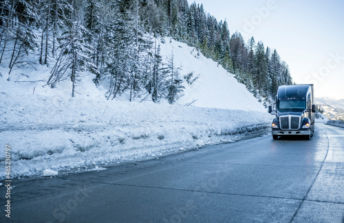Black big rig bonnet industrial semi truck transporting cargo in semi trailer running on the winding winter dangerous slippery road with snow and ice and with snowy trees on the hill side