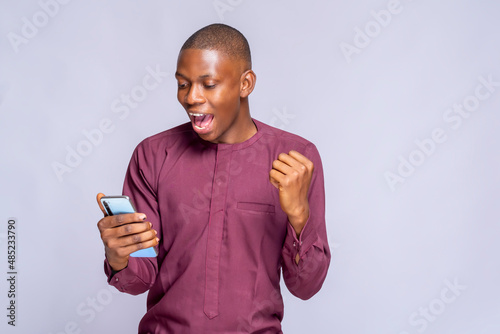 happy joyful man holds mobile phone and celebrates win makes fist bump laughs has surprised expression wears africa clothing stands sideways against white background empty space