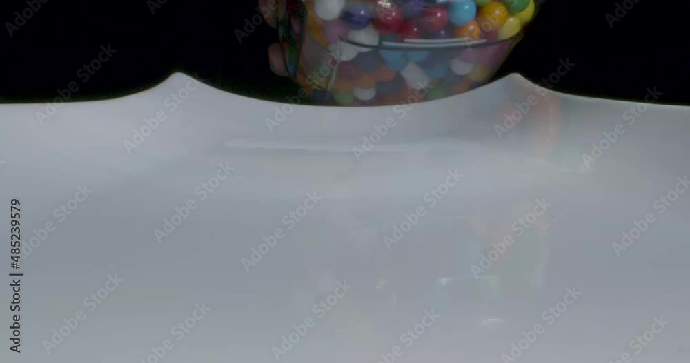 Extreme close up of colorful gumballs spilling out of a jar onto white platter in real time with audio